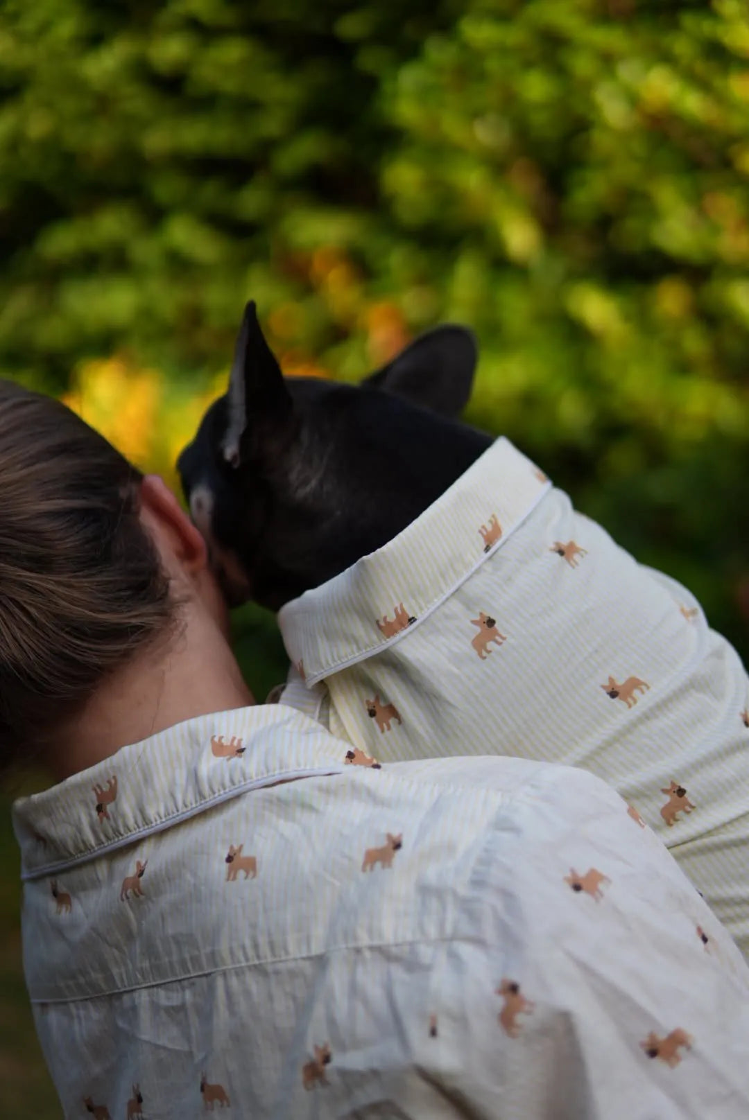 A black French Bulldog wearing a cream-colored button-up shirt with a brown dog-print pattern, while being held by a person wearing a matching shirt, against a blurred green foliage background.