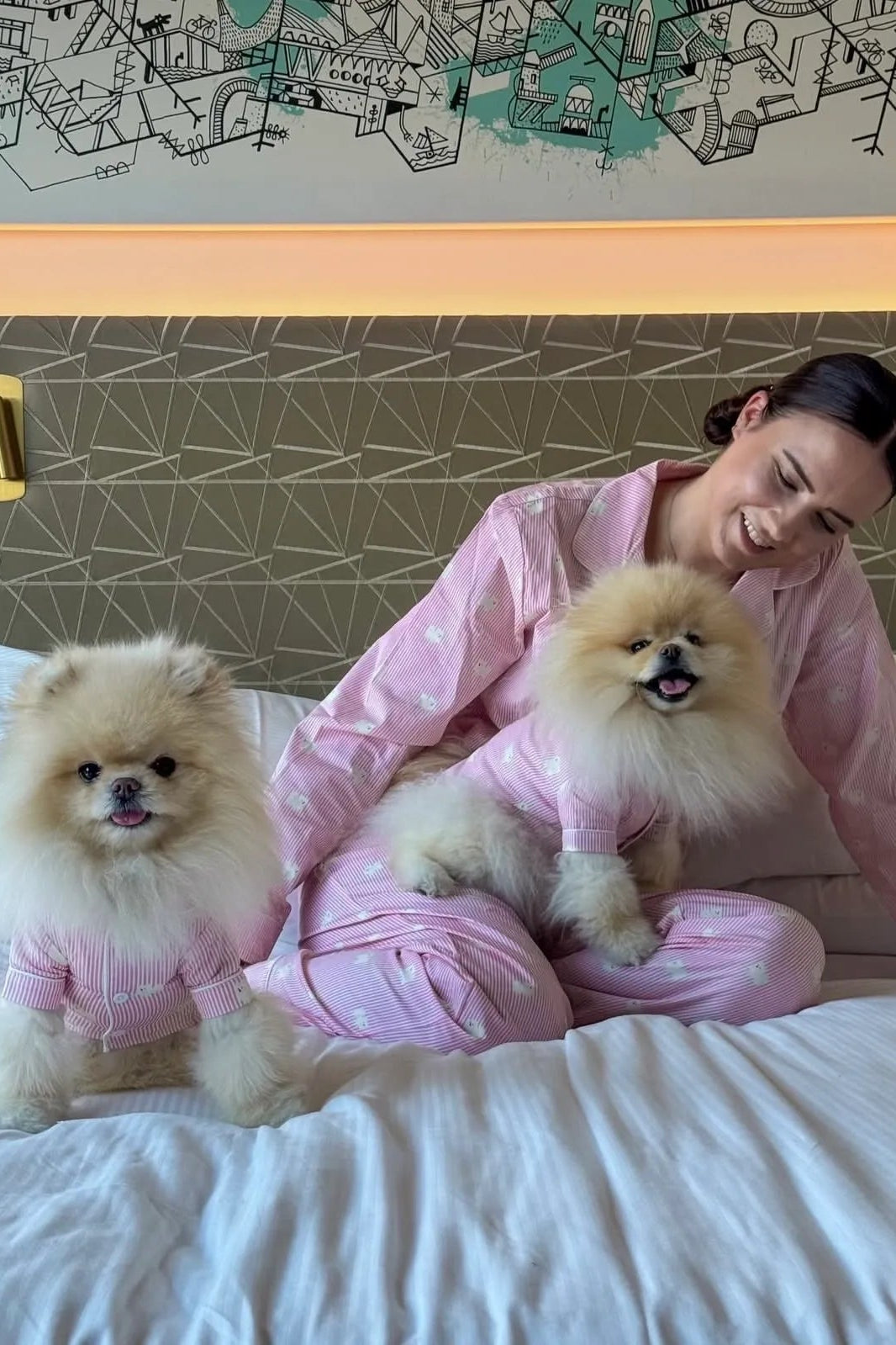 A woman and two small Pomeranian dogs wearing matching pink patterned pajamas relax on a bed in a hotel room.