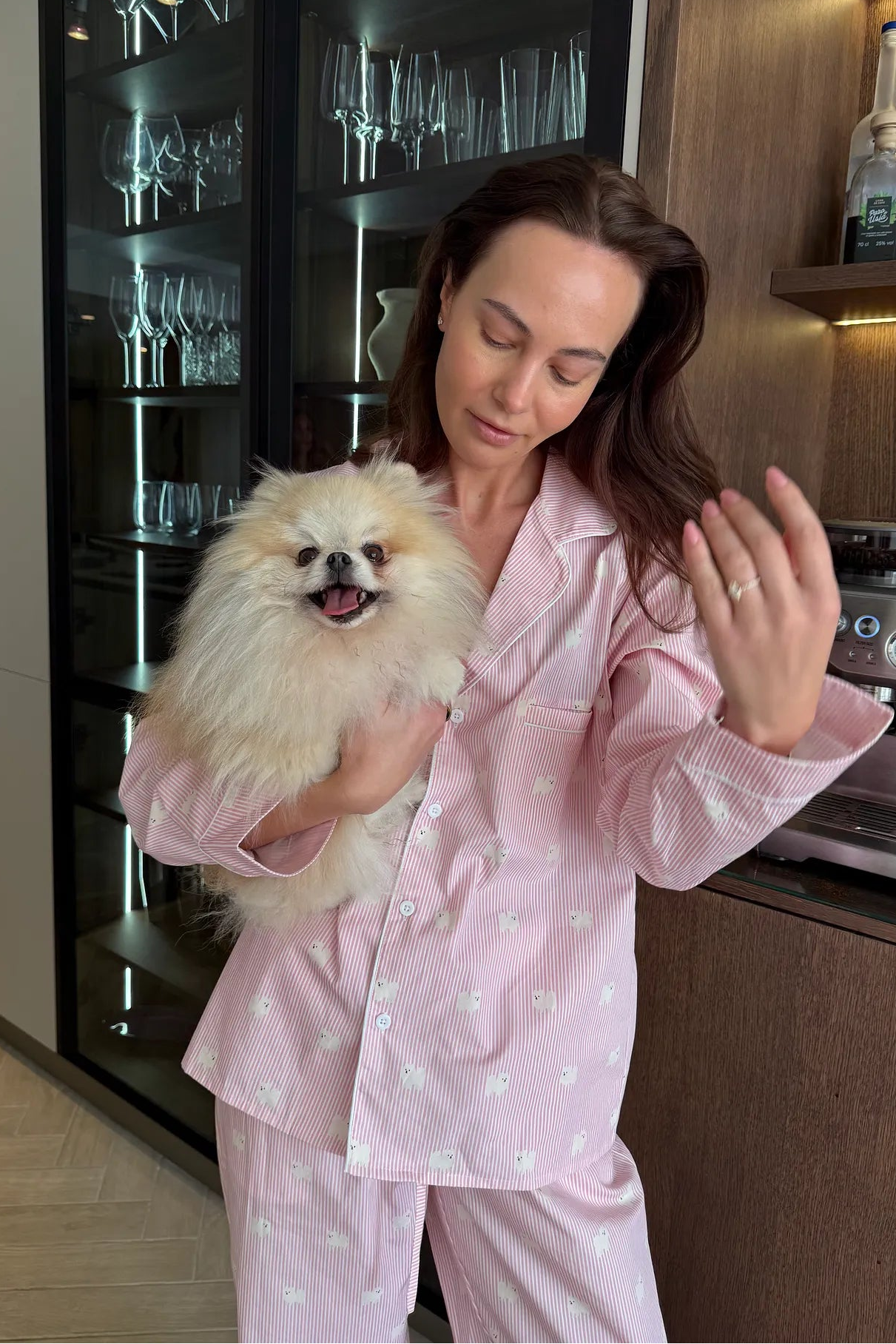 A woman in a kitchen wearing light pink pajamas with a small white floral pattern, holding a fluffy cream-colored dog.