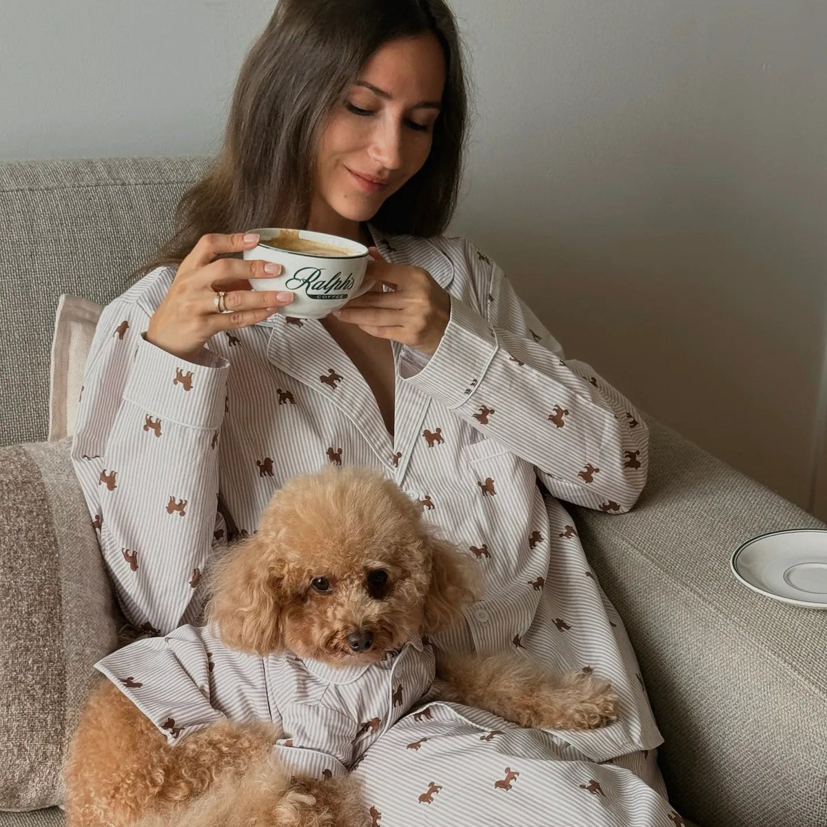 A woman and her small tan dog sit together on a light gray sofa wearing matching pajamas with a small dog motif. The woman holds a white mug.
