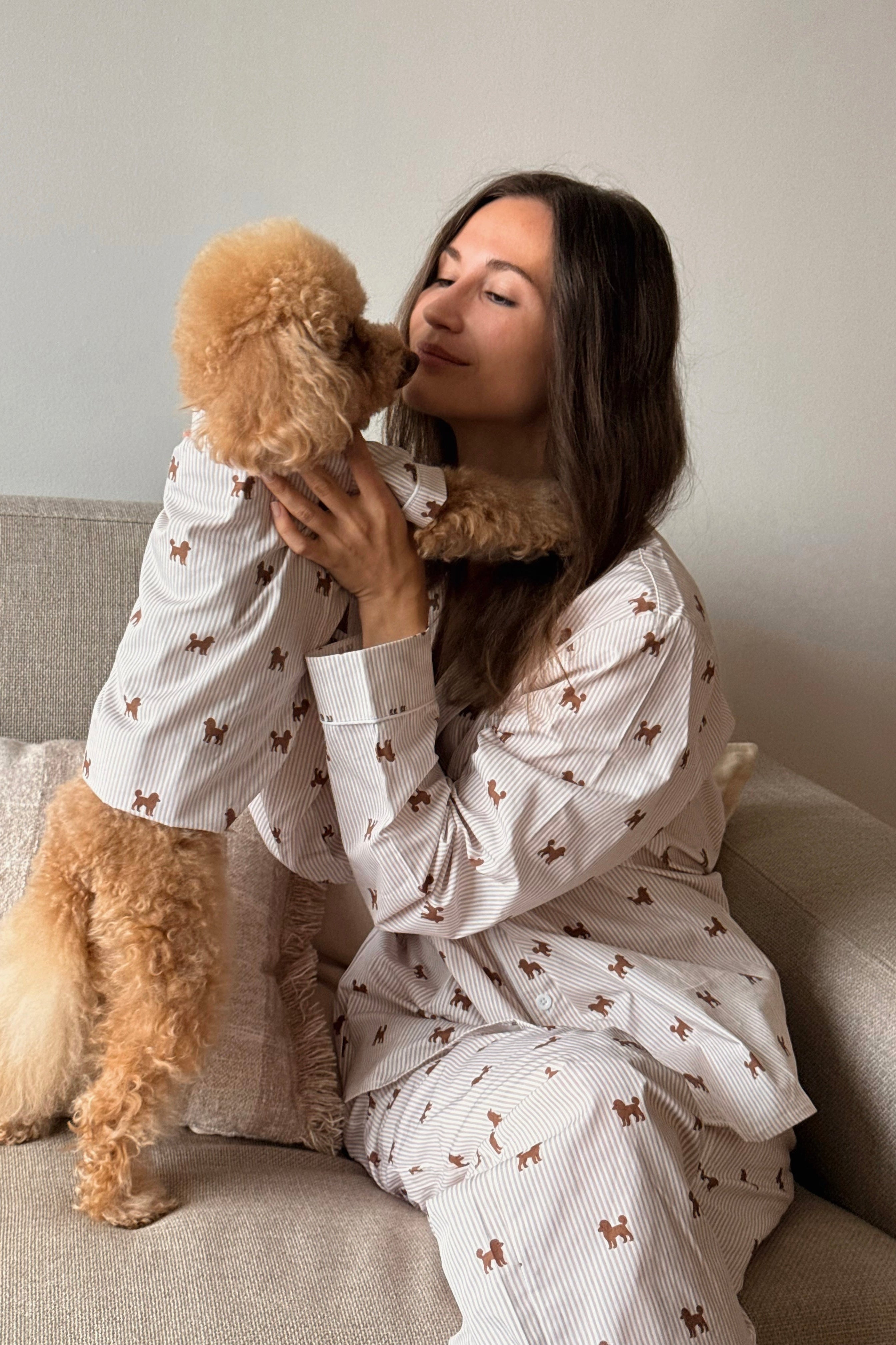 A woman wearing a matching pajama set sits on a beige couch, holding and kissing her golden poodle. The pajamas are off-white with thin vertical stripes and a repeating pattern of small brown dogs.